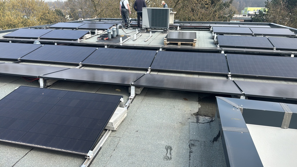 Eine Person installiert Solarpaneele auf einem grauen Flachdach unter blauem Himmel.
