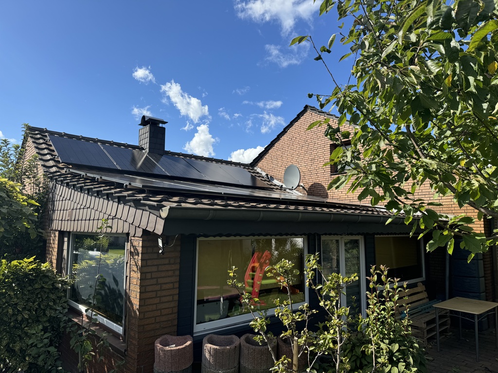 Backsteinhaus mit Solarpanels, blauer Himmel, weiße Wolken, grüne Pflanzen im Vordergrund.