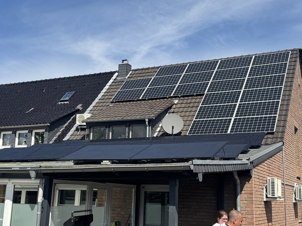 Reihenhausdach mit Solarpanels, blauer Himmel, Personen im Vordergrund, Ausdruck unbekannt.