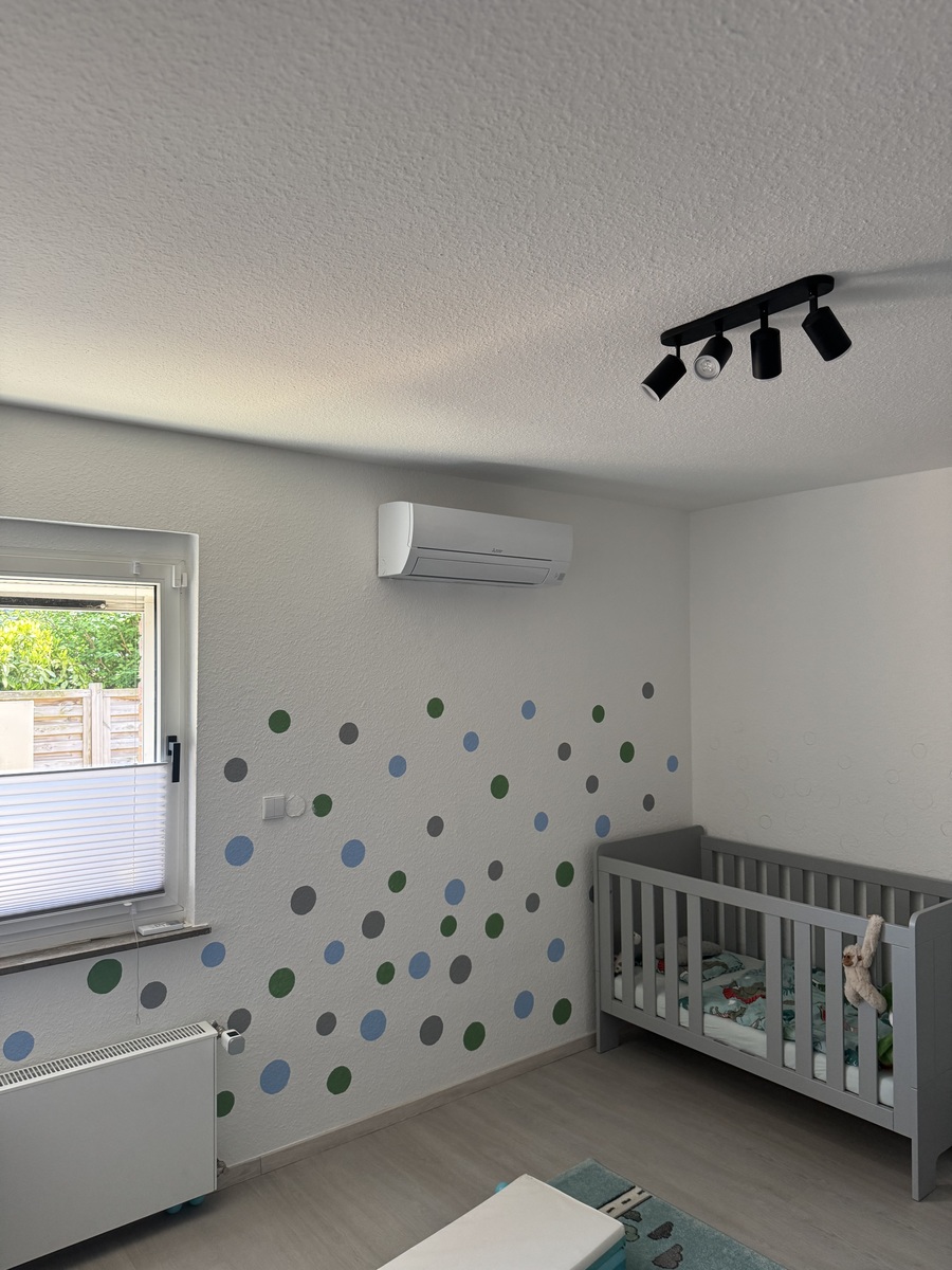 Zimmer mit grauem Babybett, gepunkteter Wand, Fenster, Klimaanlage und schwarzer Deckenleuchte.