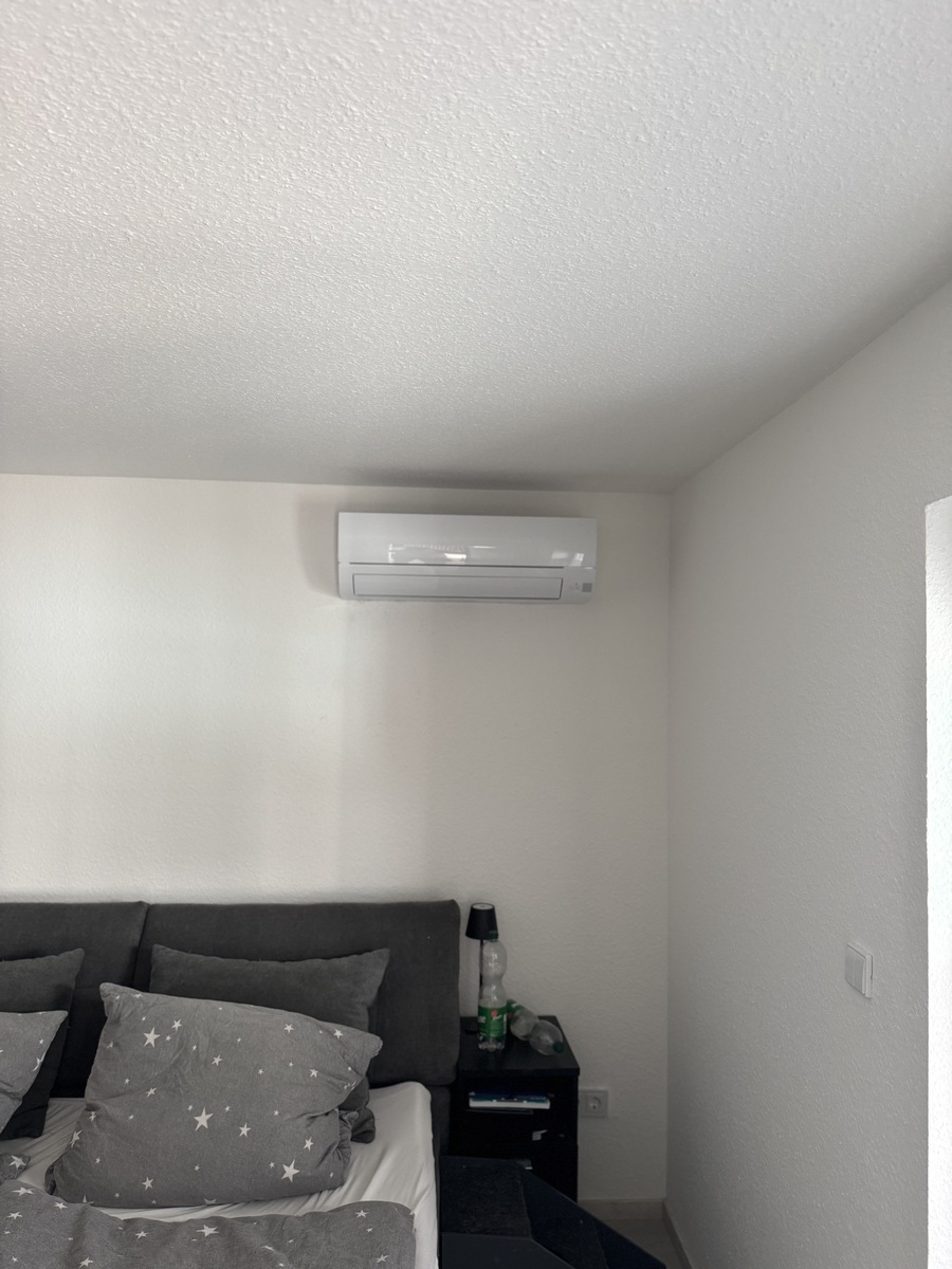 Klimaanlage an weißer Wand über grauem Bett mit sternförmigem Muster und dunklem Nachttisch.