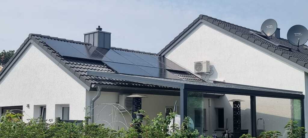 Ein weißes Haus mit schwarzem Dach und Solarpanels, daneben Sat-Schüsseln und eine grüne Hecke.